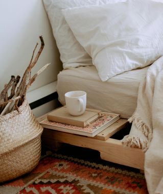 Comfortable cozy bedroom with bed wooden shelves with book and cup while wicker basket with sticks placed on ethnic styled rug